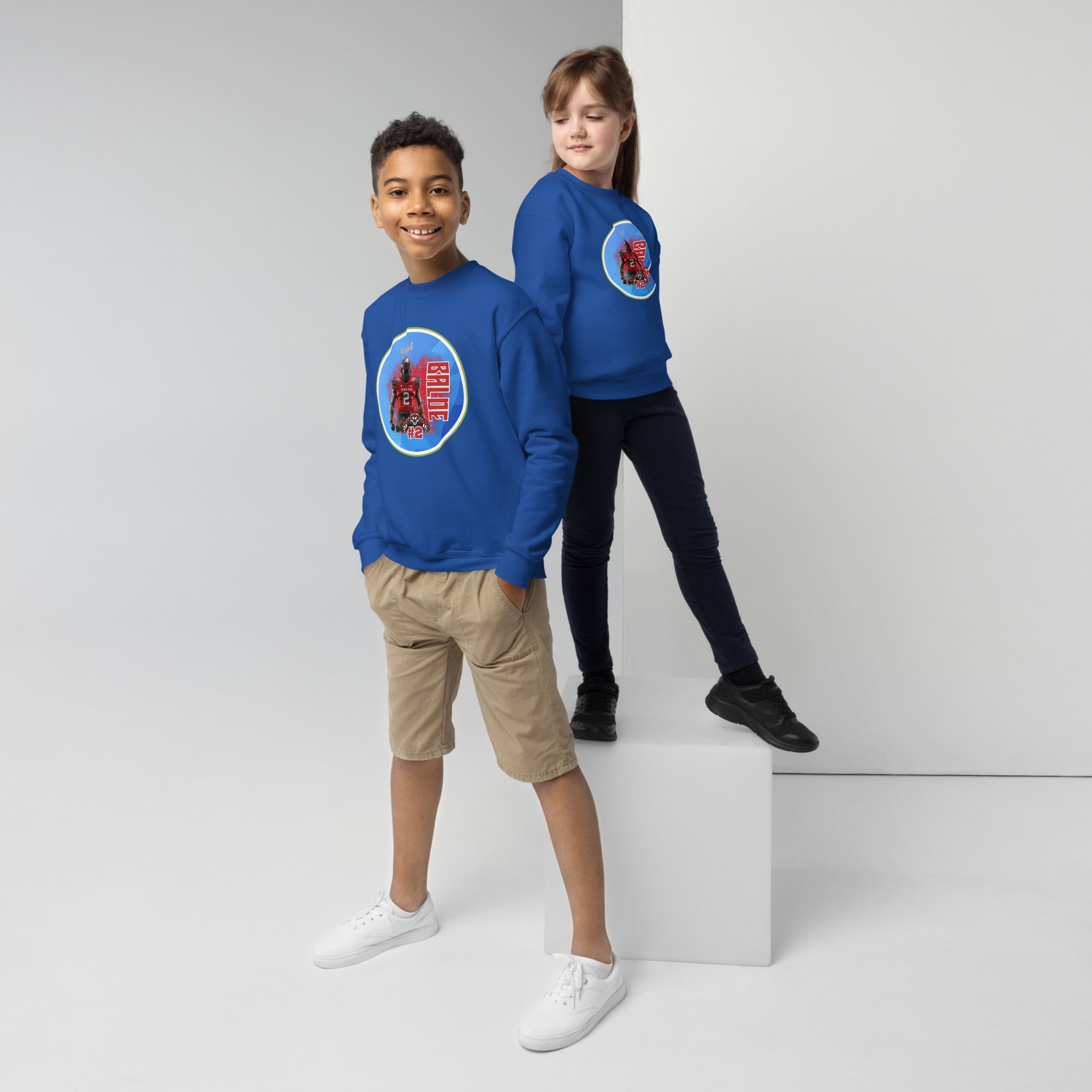 Twee kinderen dragen blauwe sweaters met een kleurrijk ontwerp op de borst. De jongen staat met zijn handen in zijn zakken en glimlacht, terwijl het meisje iets hoger staat en naar hem kijkt. De achtergrond is lichtgrijs en minimalistisch.