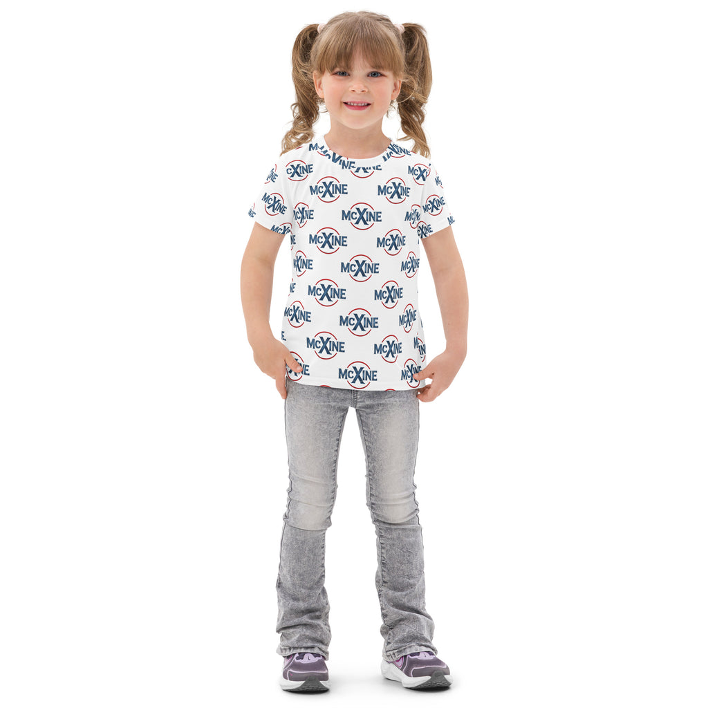 Smiling young girl wearing a white kids t-shirt with an all-over McXine logo print, paired with light grey jeans and sneakers.