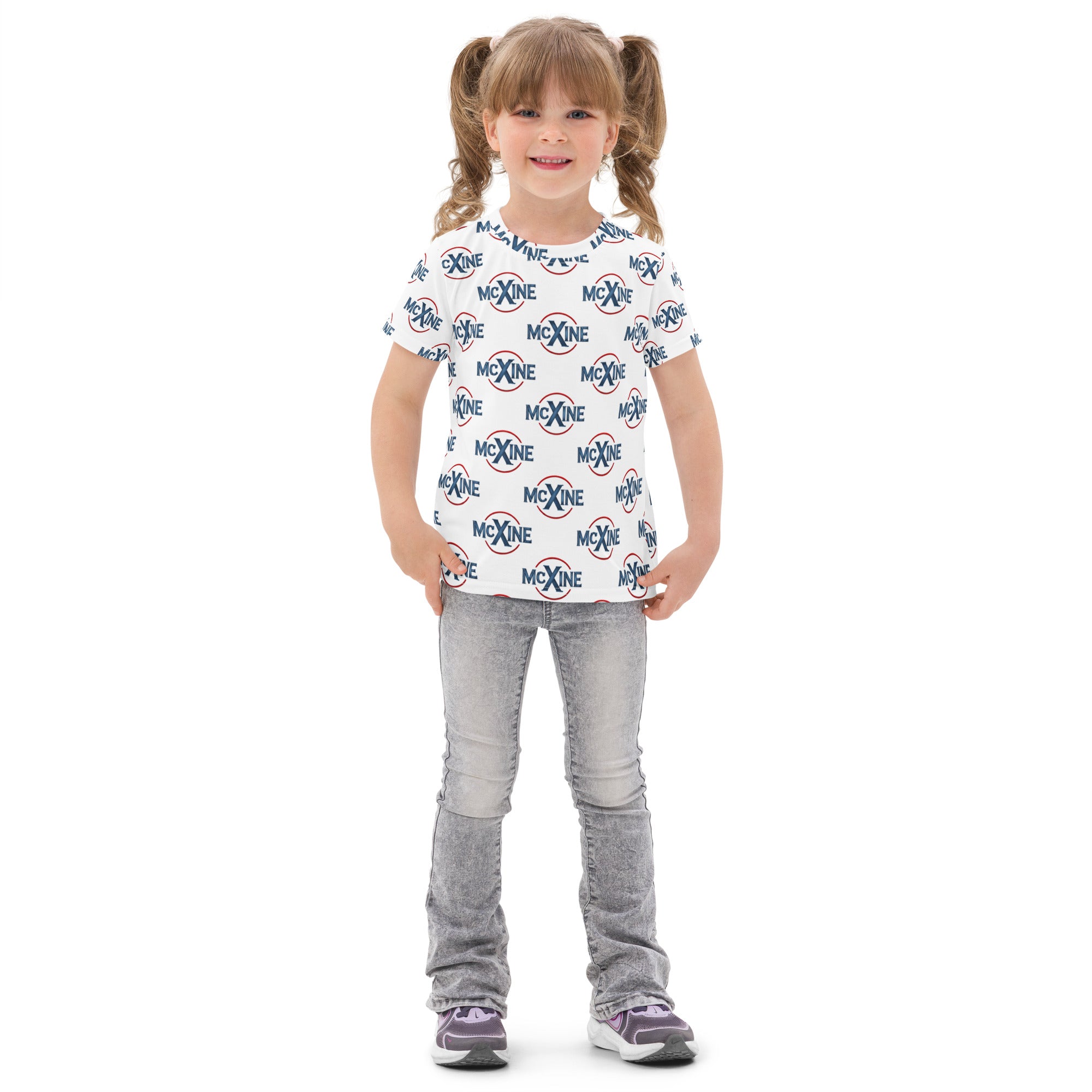 Smiling young girl wearing a white kids t-shirt with an all-over McXine logo print, paired with light grey jeans and sneakers.